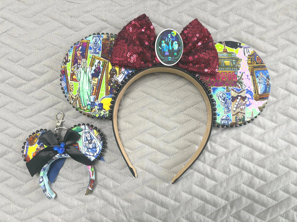 Handcrafted Haunted Manor Pixie Pair featuring a haunted attraction-inspired mouse ear headband and coordinating Mini Magic Charm with ghostly portraits, gothic manor artwork, burgundy sequin bow, black beaded trim, and handcrafted boutique styling.
