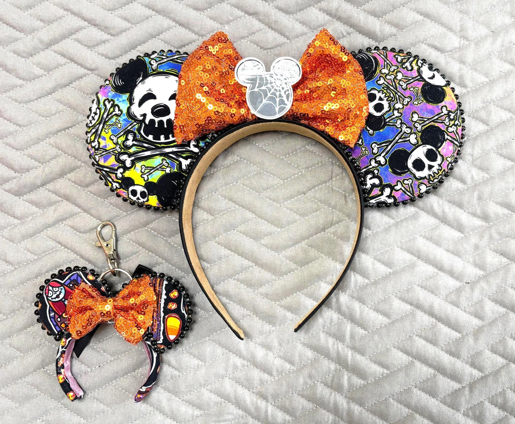 Handcrafted Spooky Park Bones Pixie Pair featuring a matching mouse ear headband and coordinating Mini Magic Charm with colorful skeleton-inspired mouse artwork, bright orange sequined bows, decorative beaded trim, and festive Halloween boutique styling.