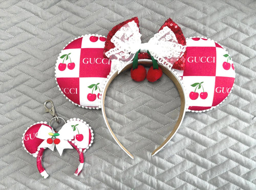 Handcrafted Cherry Belle Pixie Pair featuring a cherry-inspired mouse ear headband with ruby-red sequin bow, white lace overlay, cherry embellishments, decorative pearl-style trim, and a matching Mini Magic Charm with coordinating cherry artwork and handcrafted ribbon accents.
