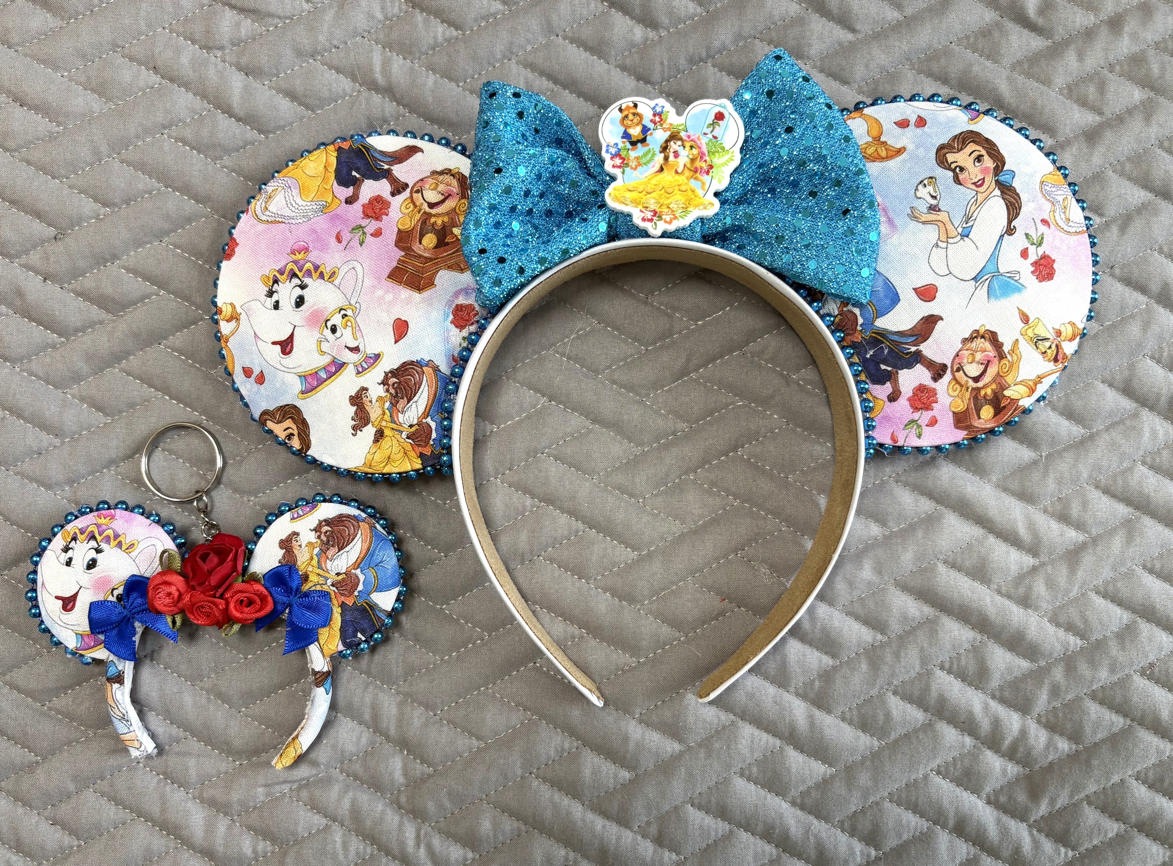Handcrafted Storybook Belle Signature mouse ear headband featuring fairy tale-inspired artwork, sapphire-blue sequin bow, coordinating princess centerpiece, decorative pearl trim, and matching Mini Magic Charm with handcrafted red roses and royal blue ribbon accents.