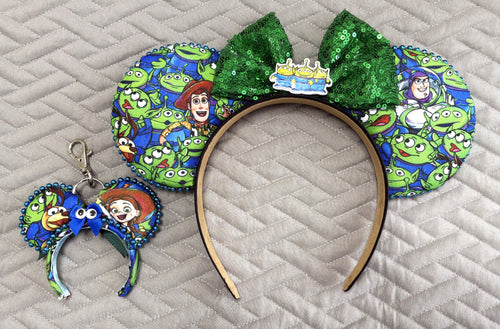 Handcrafted Cosmic Claw Pixie Pair featuring a toy-inspired mouse ear headband with playful alien and toy character artwork, emerald-green sequin bow, alien centerpiece, decorative metallic bead trim, and matching Mini Magic Charm with handcrafted alien-inspired ribbon bow.