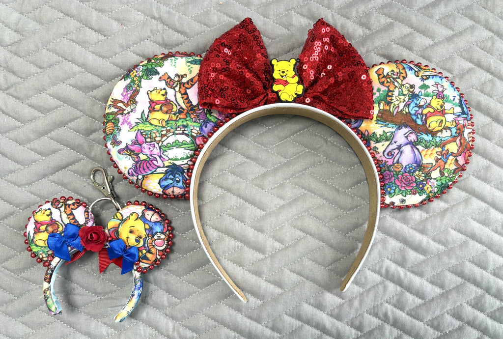 Handcrafted Hundred Acre Adventure Pixie Pair featuring a storybook-inspired mouse ear headband with woodland character artwork, ruby-red sequin bow, honey bear centerpiece, decorative metallic bead trim, and matching Mini Magic Charm with coordinating artwork and handcrafted ribbon and floral accents.