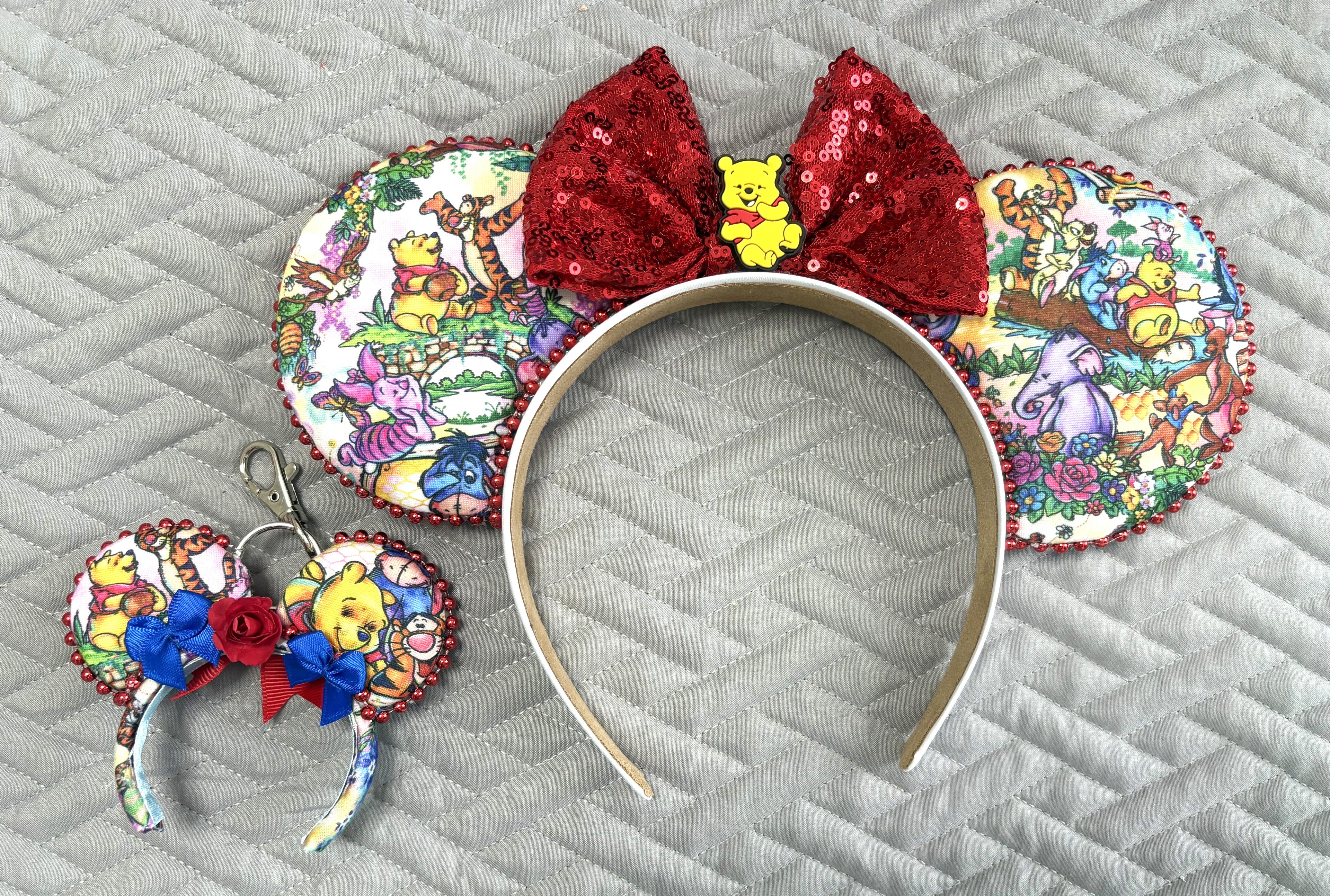 Handcrafted Hundred Acre Adventure Pixie Pair featuring a storybook-inspired mouse ear headband with woodland character artwork, ruby-red sequin bow, honey bear centerpiece, decorative metallic bead trim, and matching Mini Magic Charm with coordinating artwork and handcrafted ribbon and floral accents.