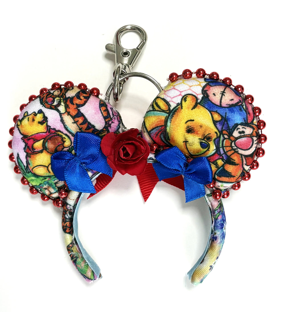 Handcrafted Hundred Acre Adventure mini mouse ear bag charm featuring storybook woodland-inspired artwork, a ruby-red bow with blue ribbon and handcrafted rose accents, decorative beaded trim, and boutique styling for handbags, backpacks, luggage, and keychains.
