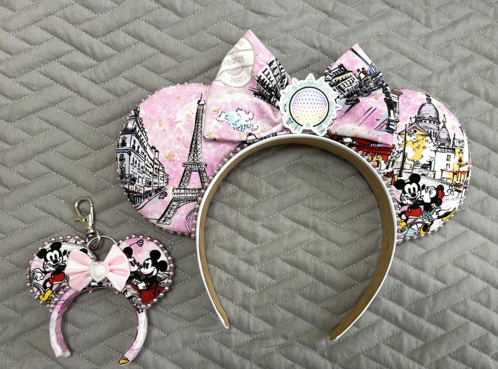 Handcrafted Bonjour Paris mini mouse ear bag charm featuring Paris-inspired artwork with the Eiffel Tower and France Pavilion styling, a blush pink boutique bow with pearl centerpiece, decorative beaded trim, and lobster clasp for handbags, backpacks, luggage, and keychains.