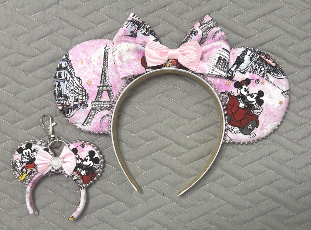 Handcrafted Parisian Elegance Pixie Pair featuring a Paris-inspired mouse ear headband with blush watercolor artwork, Eiffel Tower illustrations, romantic vintage roadster scene, elegant blush bow, decorative pearl-style trim, and matching Mini Magic Charm with coordinating artwork and boutique ribbon embellishment.