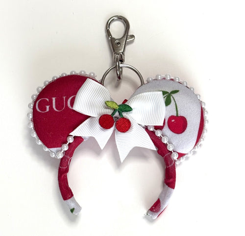 Handcrafted Cherry Belle mini mouse ear bag charm featuring cherry-inspired boutique artwork, decorative pearl trim, a layered red satin bow with floral center embellishment, and a lobster clasp for attaching to handbags, backpacks, luggage, or keychains.