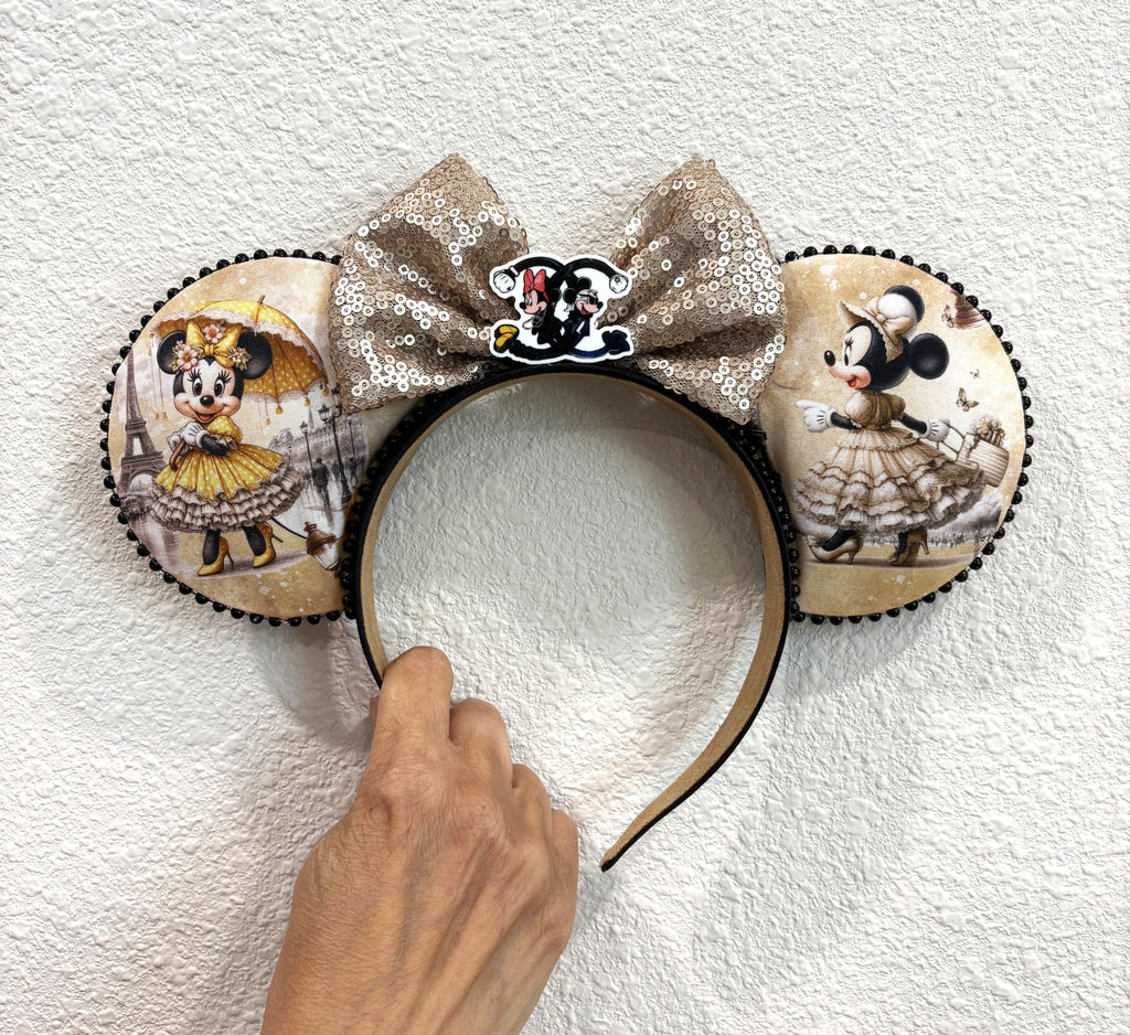 Handcrafted Parisian Elegance Pixie Pair featuring a Paris-inspired mouse ear headband and coordinating Mini Magic Charm with vintage character artwork, champagne gold sequin bow, floral embellishments, decorative trim, and handcrafted boutique styling.