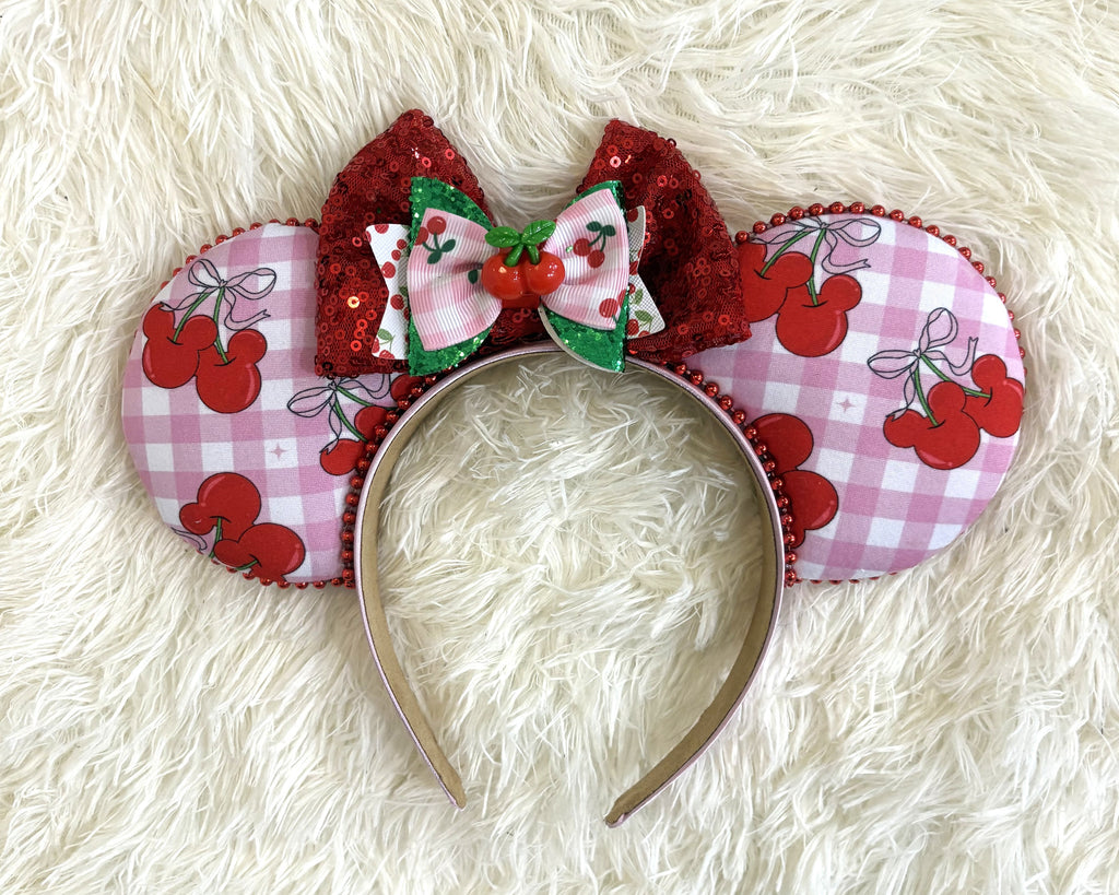 Handcrafted Cherry Jubilee Signature mouse ear headband featuring pink gingham cherry artwork, a sparkling red sequined bow, layered cherry bow accents, a cherry centerpiece, and boutique craftsmanship by Pixie Park Company.