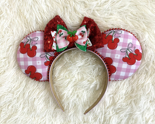 Handcrafted Cherry Jubilee Signature mouse ear headband featuring pink gingham cherry artwork, a sparkling red sequined bow, layered cherry bow accents, a cherry centerpiece, and boutique craftsmanship by Pixie Park Company.