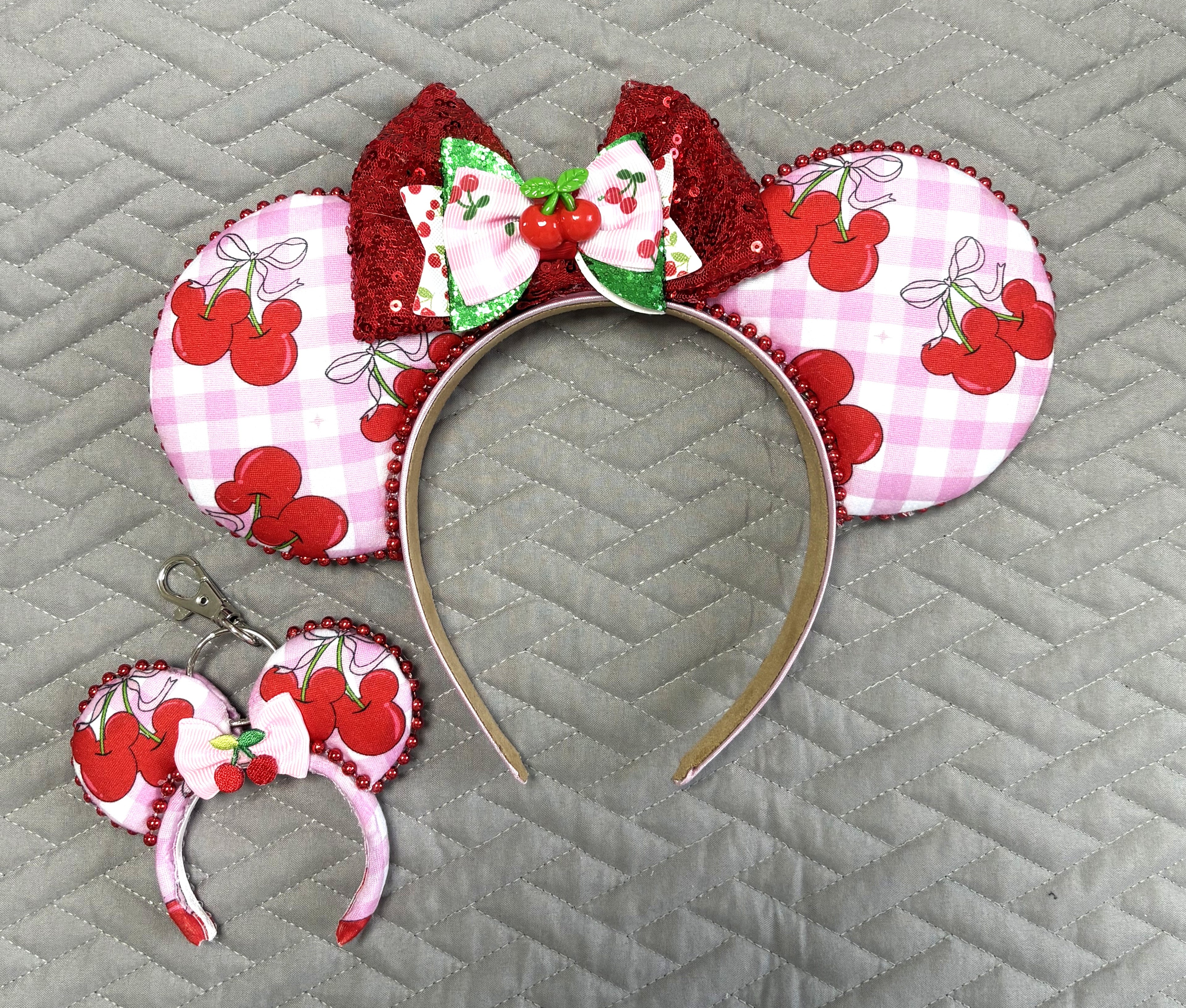 Cherry Jubilee Pixie Pair featuring a handcrafted pink gingham cherry mouse ear headband with sparkling red sequins and a coordinating Mini Magic Charm with matching cherry details.