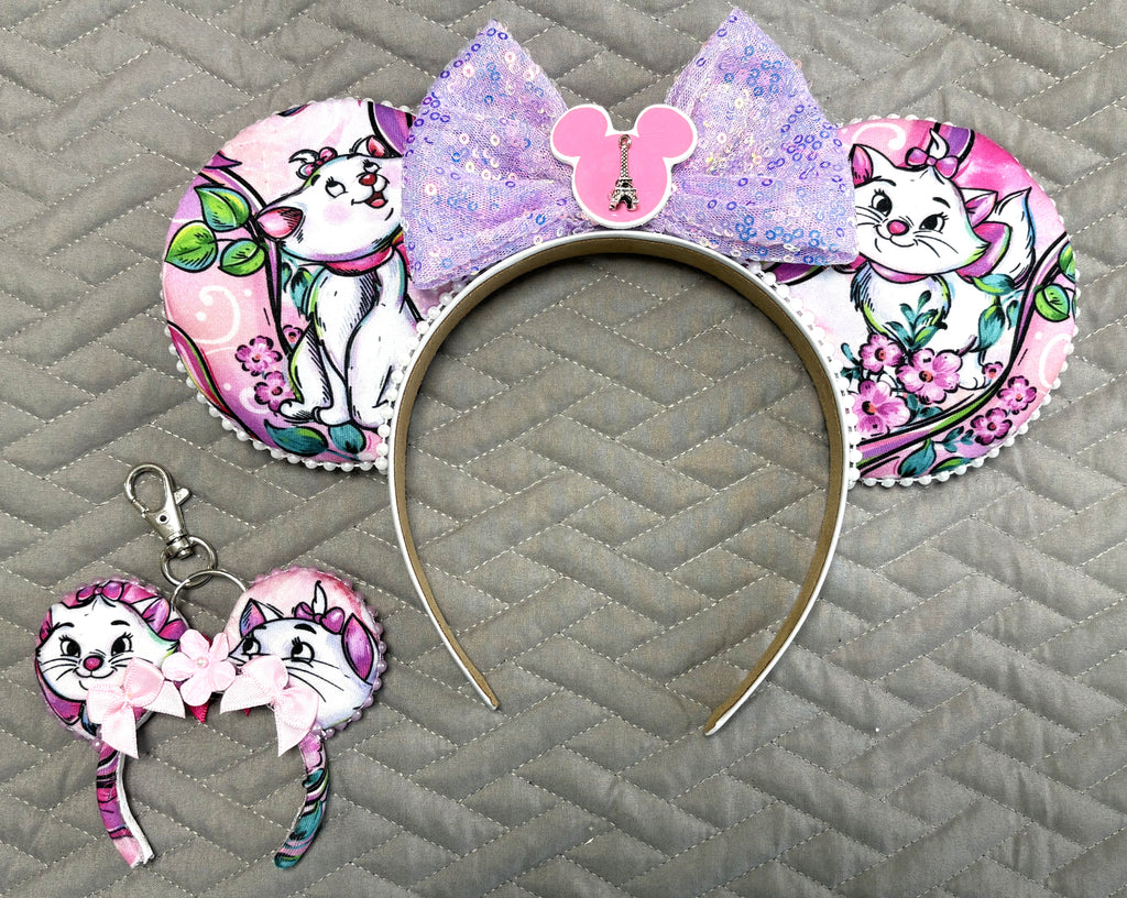 Pixie Park Company Couture Cats Pixie Pair featuring a coordinated Signature mouse ear headband and matching Mini Magic Charm with Paris-inspired pastel kitten artwork, a shimmering lavender sequin bow, decorative white beaded trim, and handcrafted boutique styling.