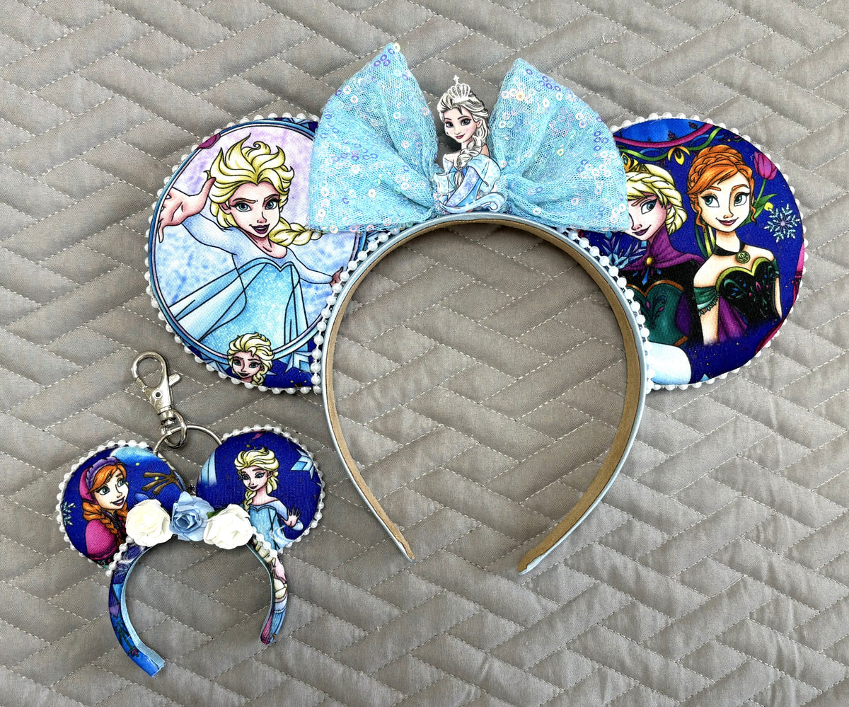 Sisters of Winter handcrafted mouse ear headband featuring two winter-princess-inspired sisters, icy blue storybook imagery, sparkling blue sequin bow and white beaded trim.