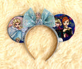 Sisters of Winter handcrafted mouse ear headband featuring two winter-princess-inspired sisters, icy blue storybook imagery, sparkling blue sequin bow and white beaded trim.