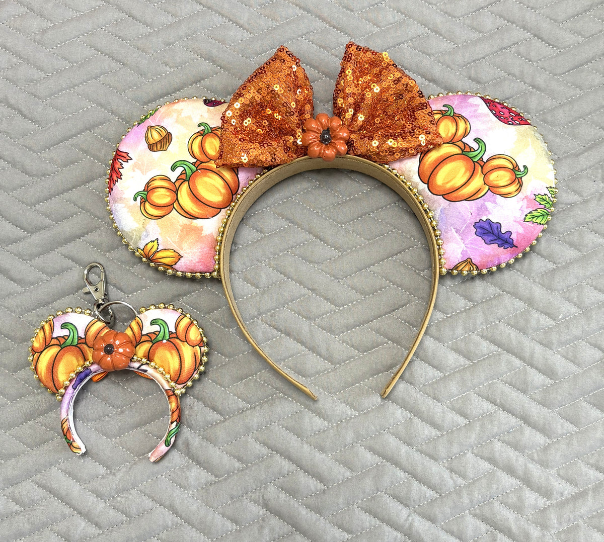 Handcrafted Pumpkin Patch Magic Pixie Pair featuring a matching autumn pumpkin-print Signature mouse ear headband with a sparkling copper-orange sequin bow, gold beaded trim, and coordinating miniature ear keychain and bag charm.