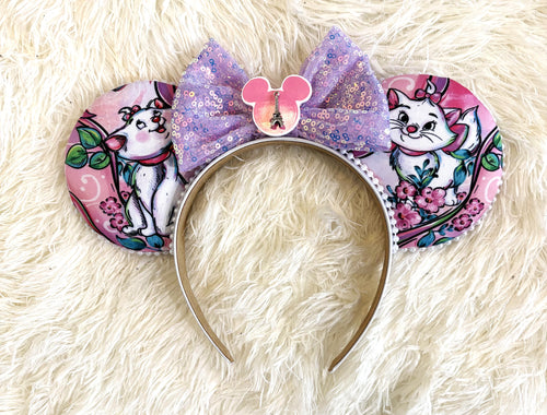 Pixie Park Company Couture Cats Signature handcrafted mouse ear headband featuring pastel Paris-inspired kitten artwork, a lavender sequin boutique bow, white beaded trim, and handcrafted boutique styling.

